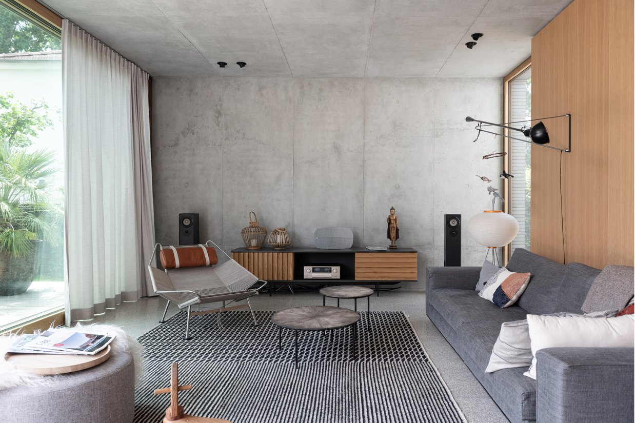 Minimalistischer Wohnraum aus Sichtbeton mit großzügiger Glasfassade, Terrazzoboden und echtholzfurnierten Einbauten aus Eiche Natur.